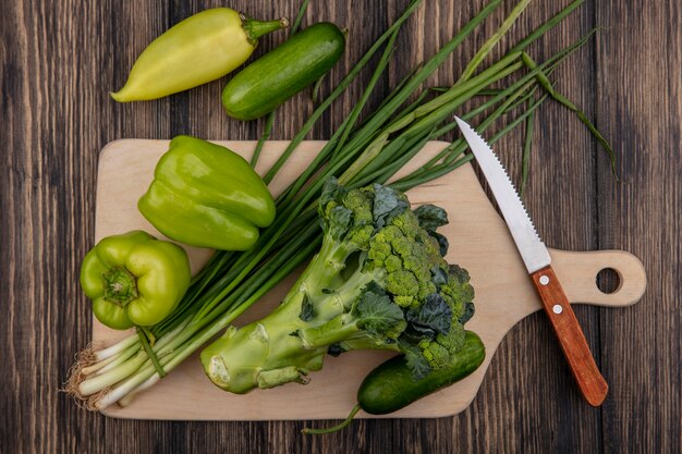 Top view cucumbers with green peppers  broccoli and green onions on a cutting board with a knife  on a wooden background