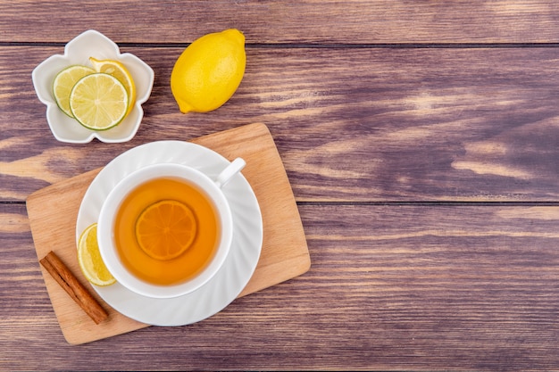 Top view of a cup of black tea with lemonnd cinnamon stick on wooden kitchen board with lemon slices on white bowl on wood