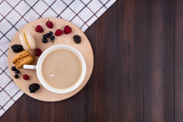 Top view of cup of cappuccino with raspberries blackberries black currants and macarons on a stand on a wooden surface