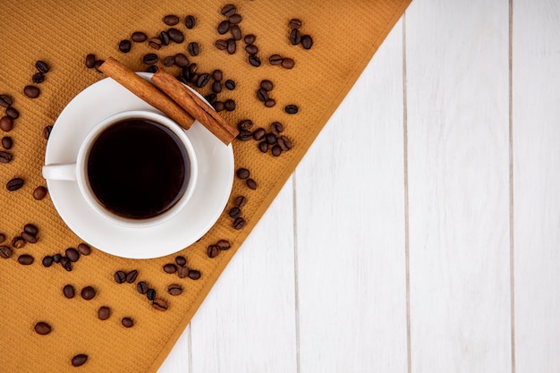 Top view of a cup of coffee on a cloth with cinnamon sticks with coffee beans on a white wooden background with copy space