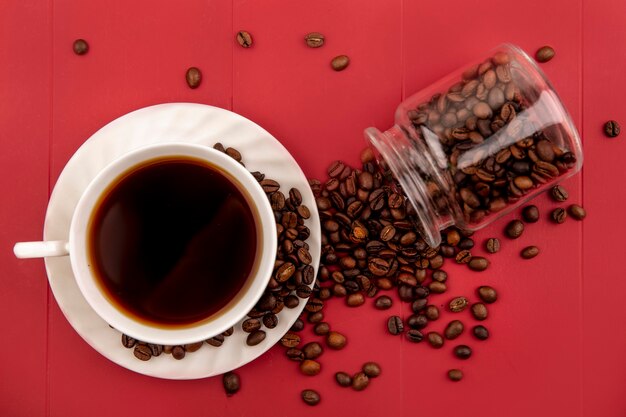 赤い背景の上のガラスの瓶から落ちてくるコーヒー豆とコーヒーのカップの上から見る