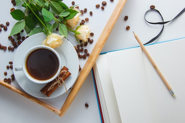 Vista superiore una tazza di caffè con i fiori, i chicchi di caffè, la matita ed il taccuino sull'orizzontale di superficie di bianco