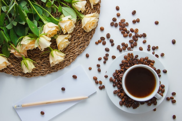 Free photo top view a cup of coffee with flowers on a trivet, coffee beans, pencil and paper on white surface. horizontal