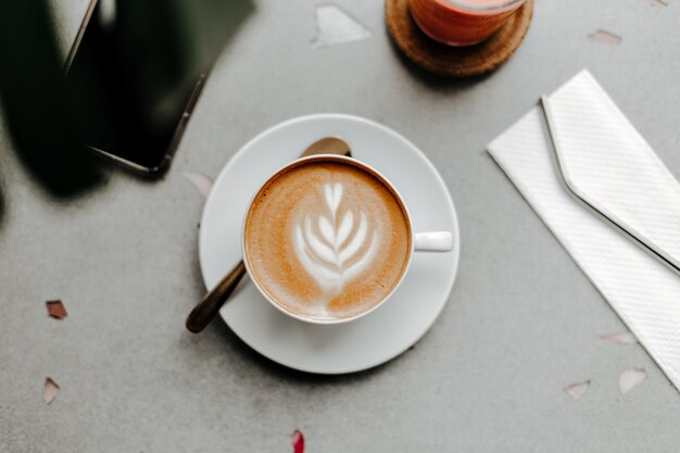 Top view of cup of coffee with foam and cream, plastic straw on napkin and telephone on marble light table