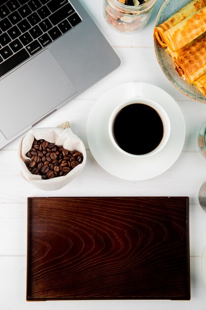 Top view of a cup of coffee with wafer rolls coffee beans in a sack laptop and a wooden board on white background