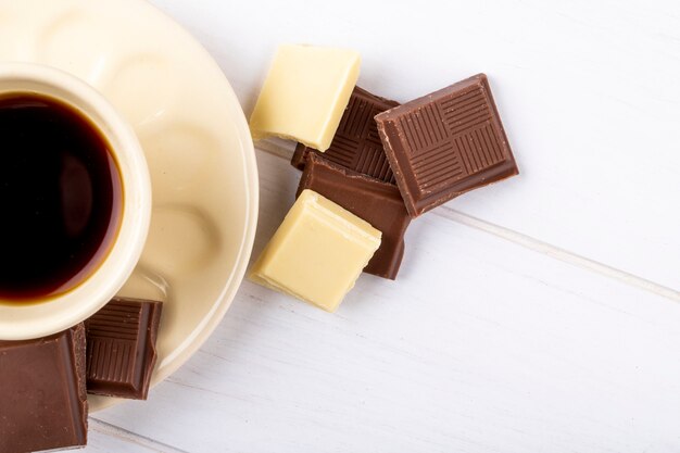 Top view of a cup of coffee with white and dark chocolate on white wooden background