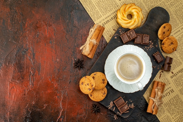 Top view of cup of coffee on wooden cutting board on an old newspaper cookies cinnamon limes chocolate bars on the left side on dark background