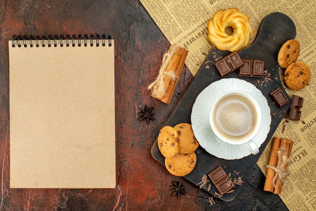Top view of cup of coffee on wooden cutting board on an old newspaper cookies cinnamon limes chocolate bars notebook on dark surface