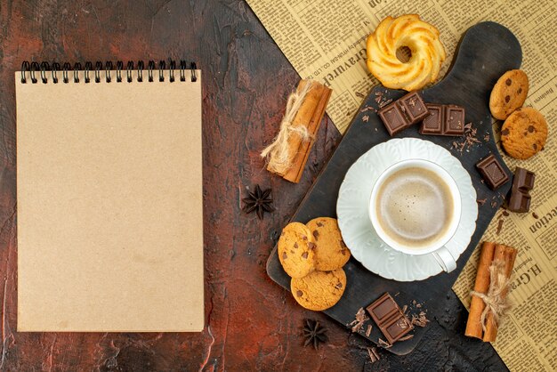 古い新聞のクッキーシナモンライムチョコレートバーノートブックの暗い表面の木製まな板上のコーヒーのカップの上面図