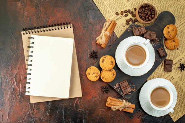 Вид сверху чашки кофе на деревянной разделочной доске на старой газете, печенье, корица, лайм, шоколадные батончики, спиральная тетрадь