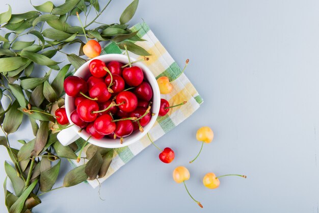 Top view of cup full of red cherries on left side and white surface decorated with leaves with copy space