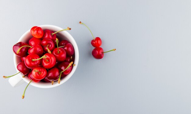 Top view of cup full of red cherries on left side and white surface with copy space