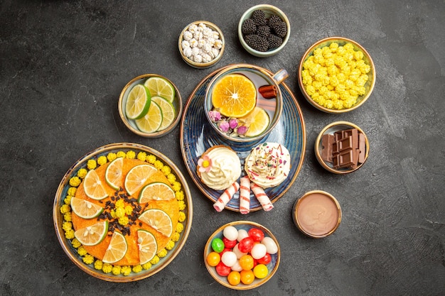 Top view a cup of herbal tea blue plate of cupcakes with cream a cup of herbal tea and sweets next to the bowls of chocolate berries citrus fruits chocolate cream and candies on the dark table