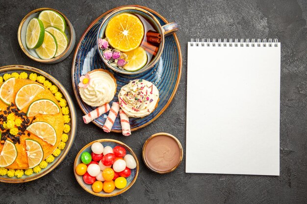 Top view a cup of herbal tea blue plate of cupcakes with cream a cup of herbal tea and sweets next to the white notebook and the bowls of citrus fruits chocolate cream and candies on the table