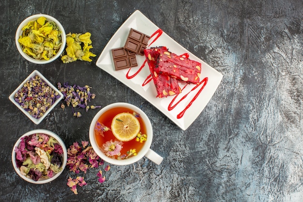 Top view of a cup of herbal tea with bowls of dry flowers and a plate of chocolate on grey ground