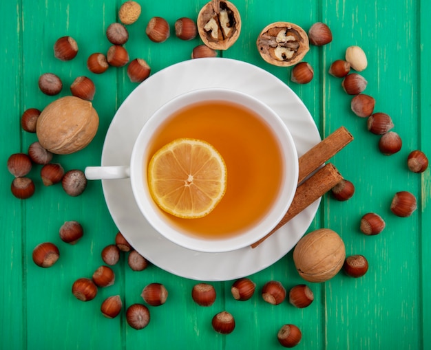 Free photo top view of cup of hot toddy with lemon inside and cinnamon on saucer with pattern of nuts and walnuts around on green background
