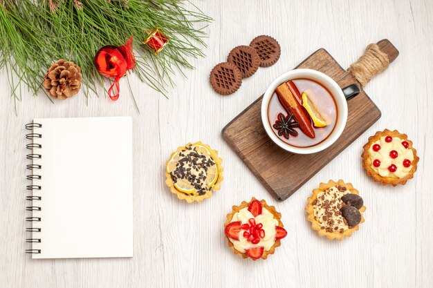 Вид сверху чашка чая с лимоном и корицей на разделочной доске, печенье и листья сосны с рождественскими игрушками и блокнотом на белой деревянной земле