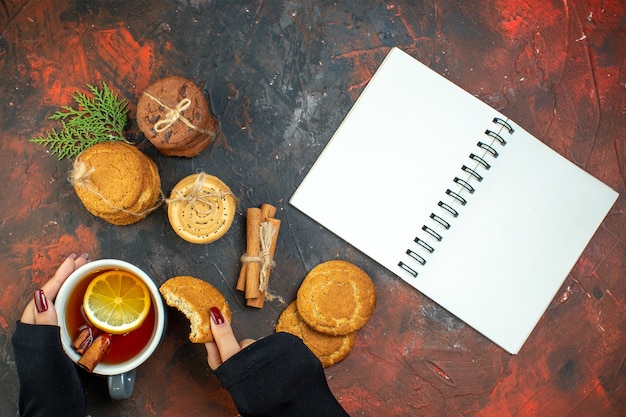 Вид сверху чашка чая в женской руке, печенье в палочках с корицей, перевязанное веревкой, блокнот на темно-красном столе Бесплатные Фотографии