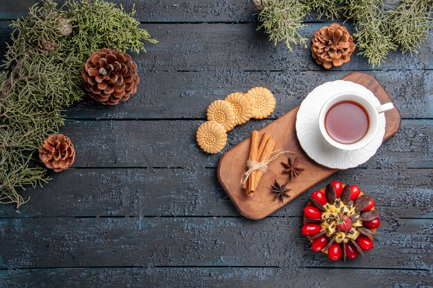 Top view a cup of tea anise seeds and cinnamon on wooden serving plate cookies pinecones berry cake on dark wooden ground