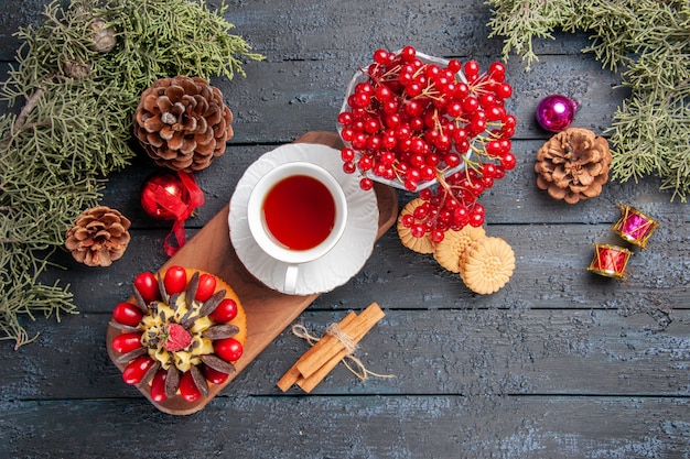 Top view a cup of tea and berry cake on wooden serving plate currant in a glass pinecones christmas toys fir-tree leaves on dark wooden table