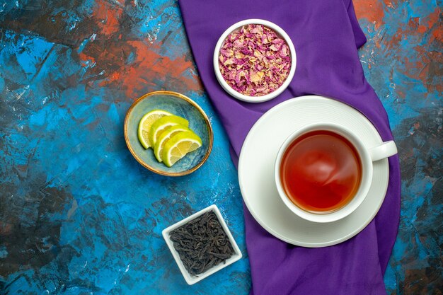 Top view a cup of tea bowls with dried flower petals and tea slices of lemon purple tablecloth on blue red surface