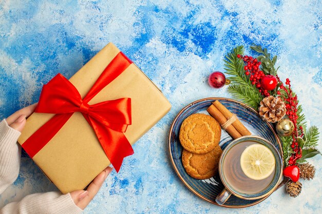 Top view cup of tea cinnamon sticks biscuits in saucer gifts in female hand on blue table