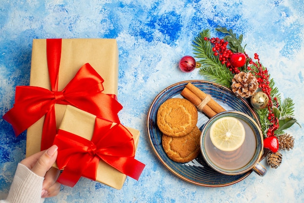 Top view cup of tea cinnamon sticks biscuits in saucer gifts in woman hand on blue table