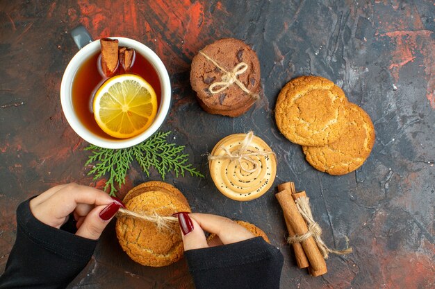 お茶のシナモンのトップビューカップは、濃い赤のテーブルに女性の手でロープで結ばれたさまざまなクッキーを貼り付けます