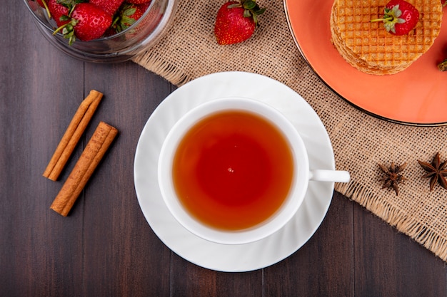 Top view of cup of tea and cinnamon with waffle biscuits and strawberries in plate and bowl on sackcloth on wood