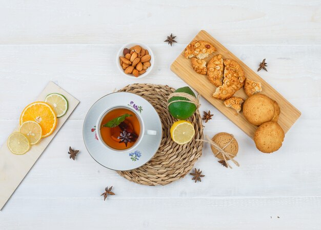 Top view of cup of tea and citrus fruits  on round placemat with cookies on a cutting board, citrus fruits and a bowl of almonds on white surface