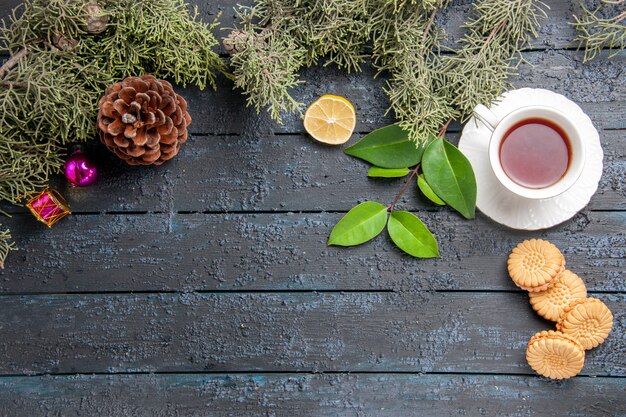 Top view a cup of tea cone fir-tree leaves christmas toys slice of lemon and biscuits on dark wooden table