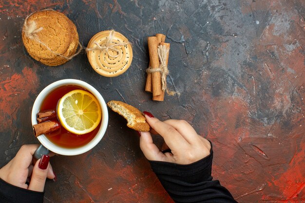 Top view cup of tea and cookie in female hand cinnamon sticks cookies tied with rope on dark red table free space