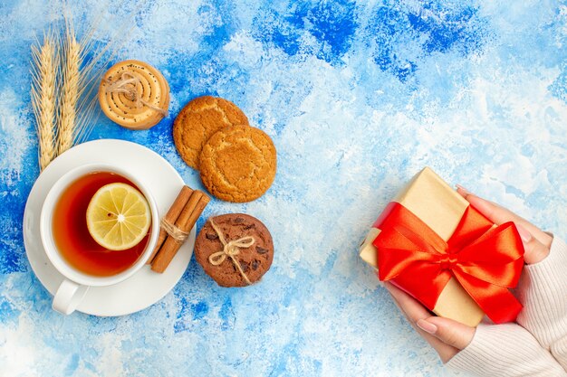 Top view cup of tea cookies gift in female hand on blue table free space