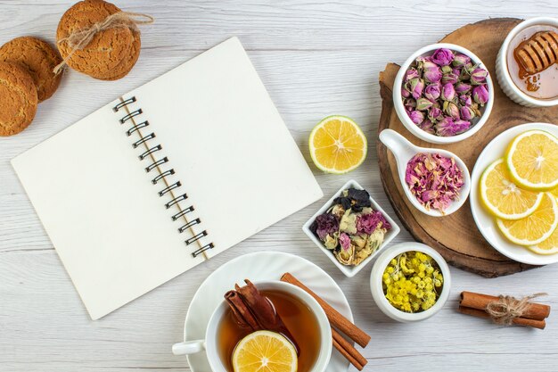 Вид сверху на чашку чая, печенье и различные травы и нарезанный лимонный мед с ложкой на деревянном подносе рядом с открытой записной книжкой на белом фоне