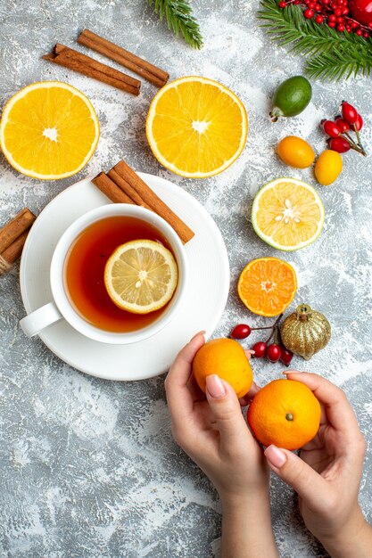 Top view a cup of tea cut lemons cinnamon sticks mandarins in female hand on grey background