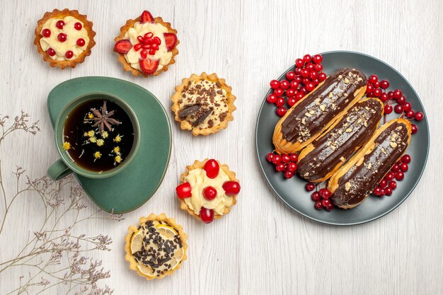 Top view a cup of tea and different cookies at the left and chocolate eclairs and currants on the grey plate at the right side of the white wooden table