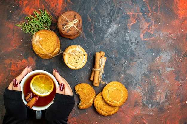 Free photo top view cup of tea in female hand cinnamon sticks cookies tied with rope on dark red table free space