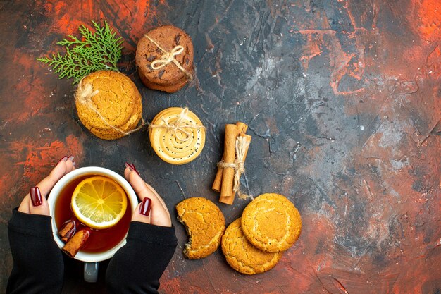 Top view cup of tea in female hand cinnamon sticks cookies tied with rope on dark red table free space