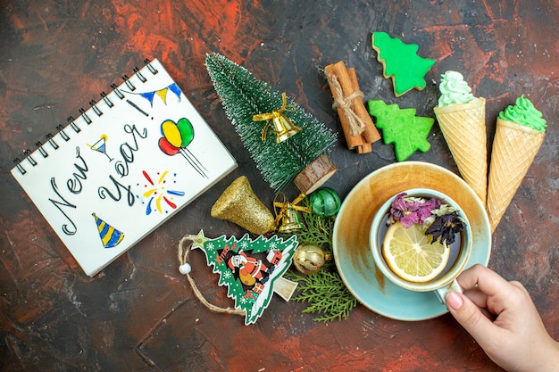 Free photo top view cup of tea in female hand ice creams cinnamon sticks xmas tree cookies xmas ornaments new year written on notebook on dark red table