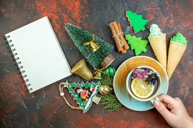 Top view cup of tea in female hand ice creams cinnamon sticks xmas tree cookies xmas ornaments notebook on dark red table