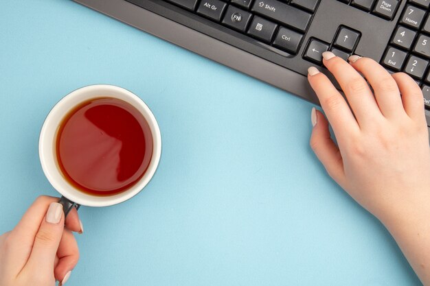 Top view cup of tea in female hand keyboard on blue table