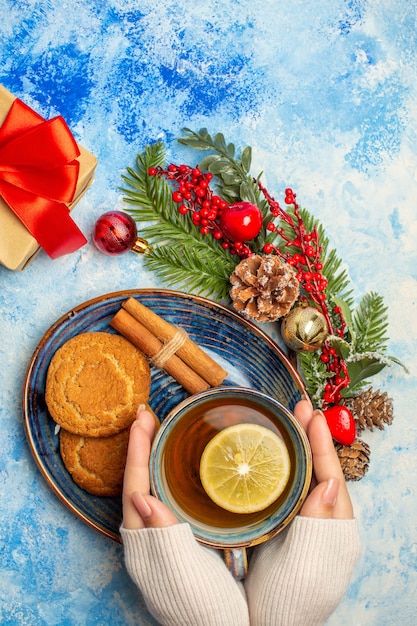 Foto gratuita vista dall'alto tazza di tè in mano femminile fette di limone bastoncini di cannella biscotti in piattino sul tavolo blu