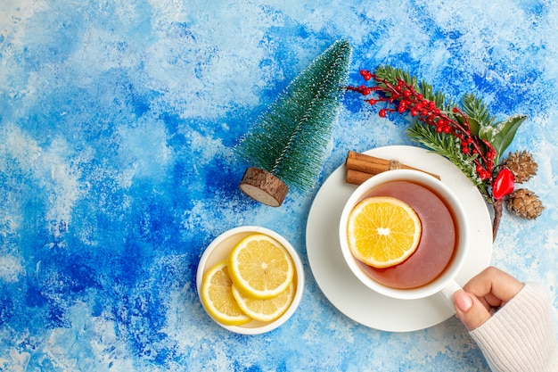 Top view cup of tea in female hand xmas tree branch cut lemons on saucer on blue table