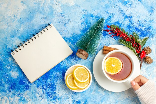 Вид сверху чашка чая в женской руке ветка рождественского дерева разрезанные лимоны на блюдце, блокнот на синем столе