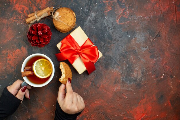 Top view cup of tea flavored by lemon and cinnamon and cookie in female hand xmas gift red pinecone cinnamon sticks cookies on dark red table with copy place
