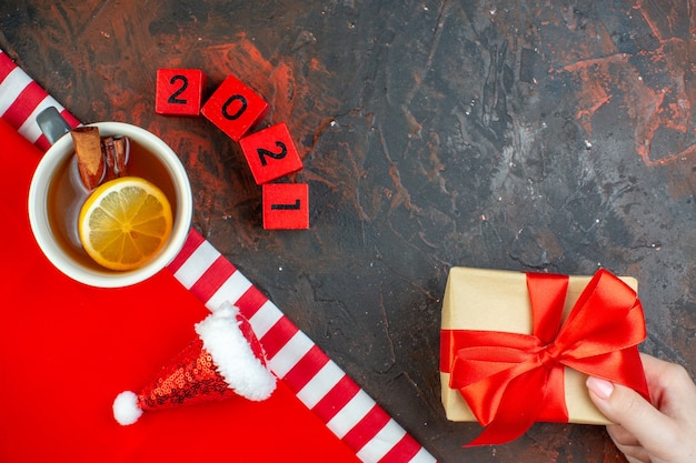 Top view cup of tea flavored by lemon and cinnamon mini santa hat on red tablecloth wood blocks gift in woman hand on dark red table free space