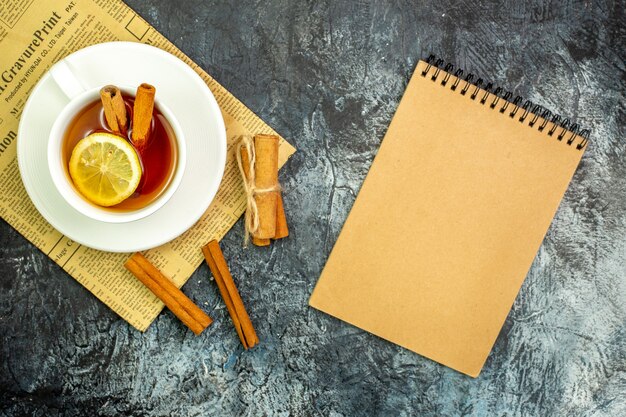 Top view cup of tea flavored by lemon and cinnamon on newspaper cinnamon sticks notebook on dark table