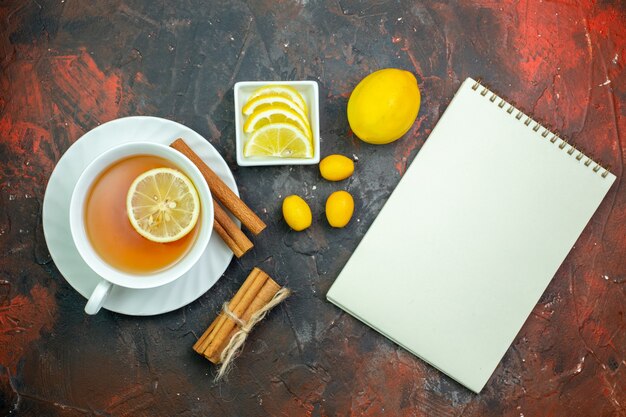 Top view cup of tea flavored by lemon cumcuat lemon slices in small bowl cinnamons notepad on dark red background