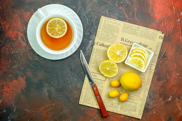 Free photo top view cup of tea flavored by lemon cumcuats lemon slices in small bowl cinnamon sticks knife on newspaper on dark red table