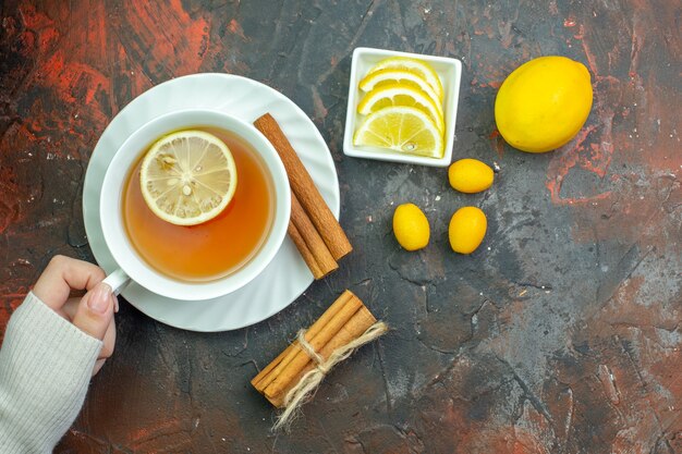 짙은 빨간색 테이블에 있는 작은 그릇 계피에 있는 여성 손 cumcuat 레몬 조각에 레몬으로 맛을 낸 차 한 잔